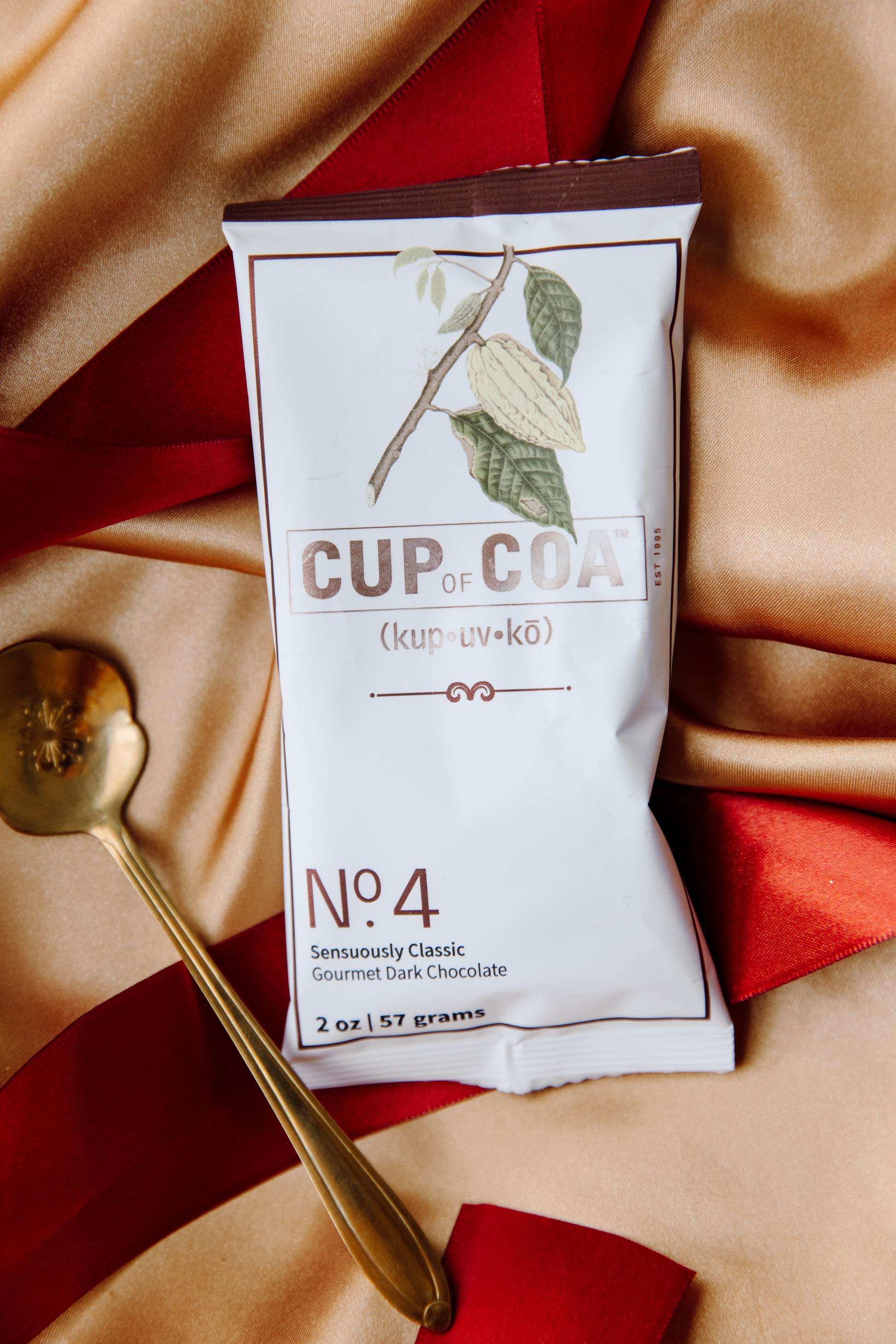 Cup of Coa No. 4 Sensuously Dark single-serve packet displayed on silk, accented with an orange ribbon and gold spoon.